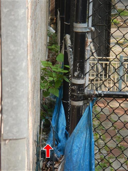 Blue tarp and debris at the base of an exterior wall beside a black pipe and chain-link fence, red arrow pointing at the debris