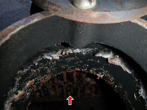 View down into an opened cast iron boiler showing a cracked fire box and heavy soot buildup, red arrow