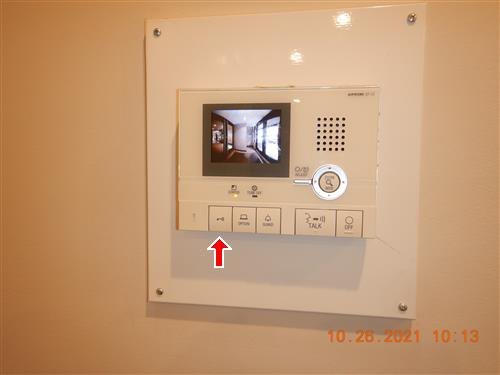 Wall-mounted video intercom panel showing a hallway on its screen, red arrow at a button, date stamp