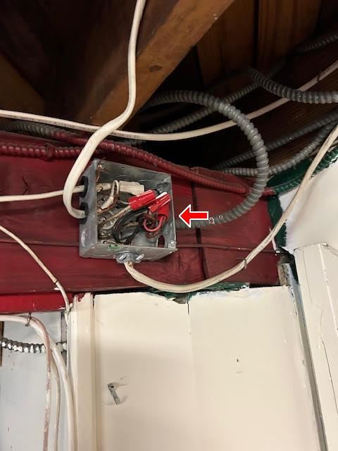 An open junction box with its cover missing, wire connectors exposed, mounted on a joist in an older basement alongside armoured cable.