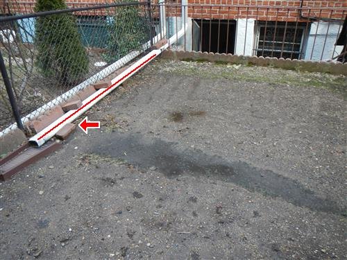 A rear yard with a downspout extension running along the fence line, marked with a red arrow.