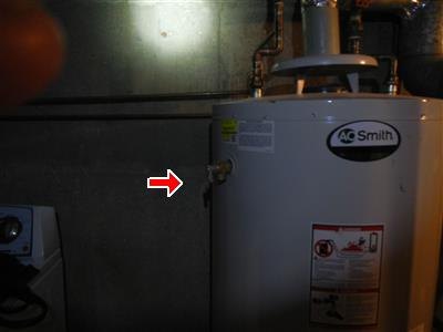 White gas water heater in a basement with a red arrow at the relief valve on its side.