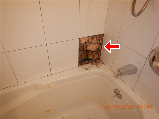 A bathtub surround with one ceramic tile removed, red arrow at the exposed wall cavity.