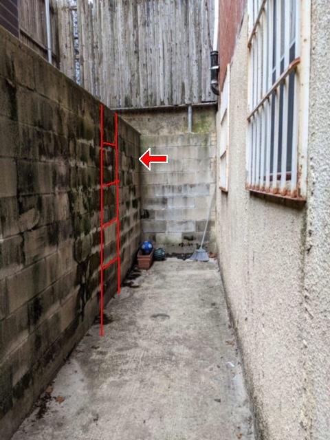 A narrow below-grade passage between buildings with a ladder outline drawn in red on the block wall.