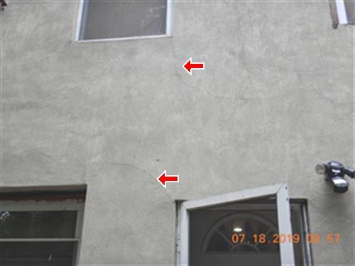 A stucco rear wall with red arrows marking cracks below a window and above the rear door.
