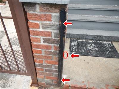 Black steel gate latch plate set into a brick pier beside a metal gate, with three red arrows and a circled hole.
