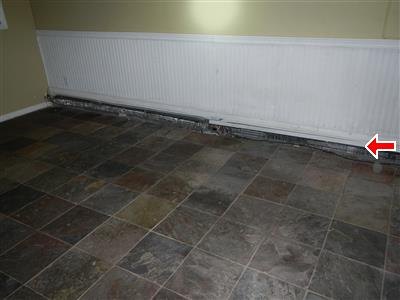 White baseboard heating unit along a wall with its lower front covers missing, red arrow at one end.