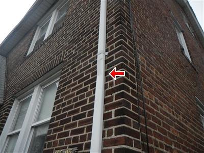 Looking up a brick wall corner at a white downspout, red arrow at a crushed section of the pipe.