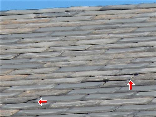 Sloped slate roof with two red arrows at broken and slipped pieces.