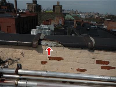 A rooftop parapet with a broken ceramic coping section marked by a red arrow, painted brick spalling below.