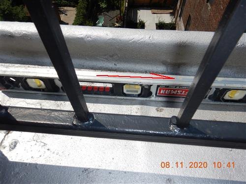 A spirit level lying in a silver-coated built-in gutter at a roof edge, seen through an iron railing, with a red line showing the slope.
