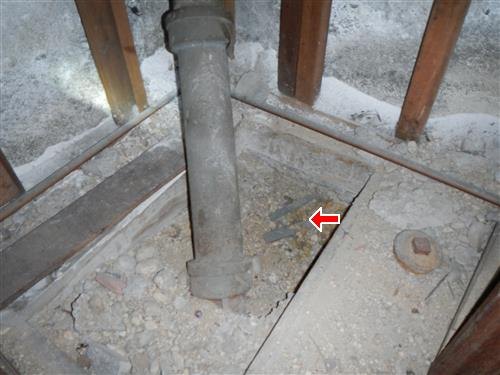 A cast iron drain pipe angling into a rough unfinished floor, with a red arrow at its base