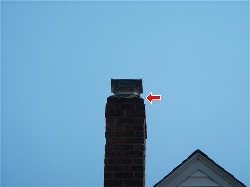 A brick chimney against a clear sky with a red arrow at the cement cap below the metal cap and screen