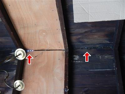 Underside of a garage roof at the peak, plywood panel beside dark wood boards, two red arrows pointing at daylight through an opening at the ridge