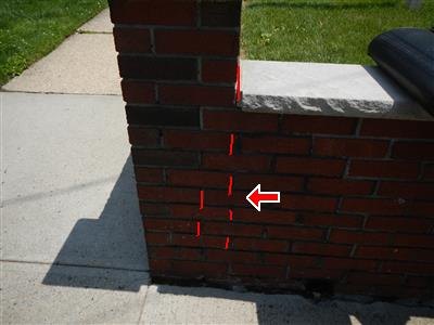 A low brick wall with a stone cap beside a sidewalk, a crack traced in red down the mortar joints, red arrow