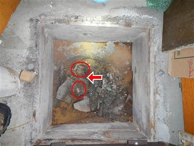 A square pit in a basement floor with the whole-house sewer trap circled in red under debris
