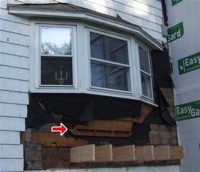Bay window with siding removed below, exposing black felt, sheathing and new support lumber, with a red arrow at the framing