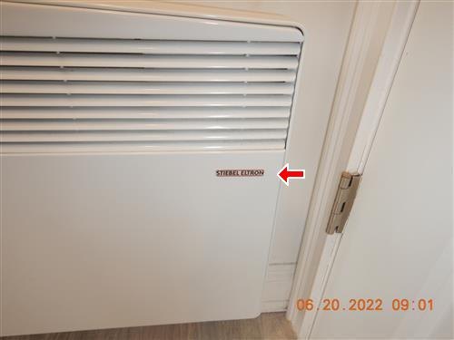 A wall-mounted electric convector heater beside a door frame, with an arrow at its brand label