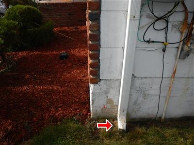 A downspout ending at a foundation corner with no splash block, red arrow at the discharge point