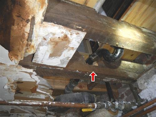 Old wood cellar joists overhead with a drain pipe passing through a cut double joist, red arrow at the cut