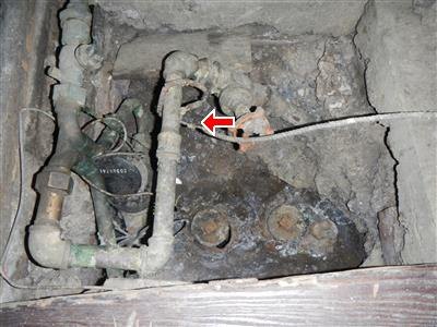 Corroded water piping and valves in a dirt-floored access pit, with a red arrow at the broken ground wire clamp on the water main